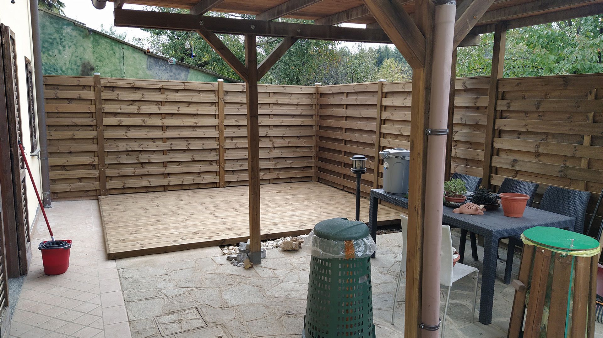 Patio coperto con pali di legno, angolo recintato, pavimento piastrellato, tavolini e vasi di piante.