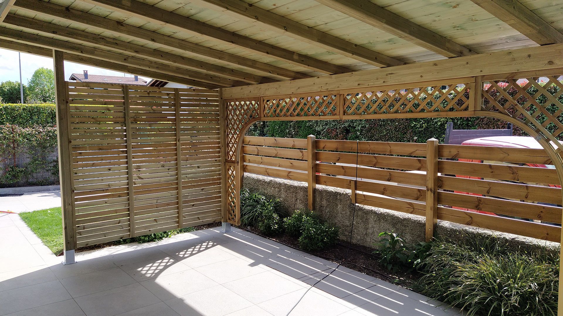 Patio coperto con pareti a traliccio in legno e recinzione a doghe, adiacente a un pavimento in cemento e a una siepe da giardino.