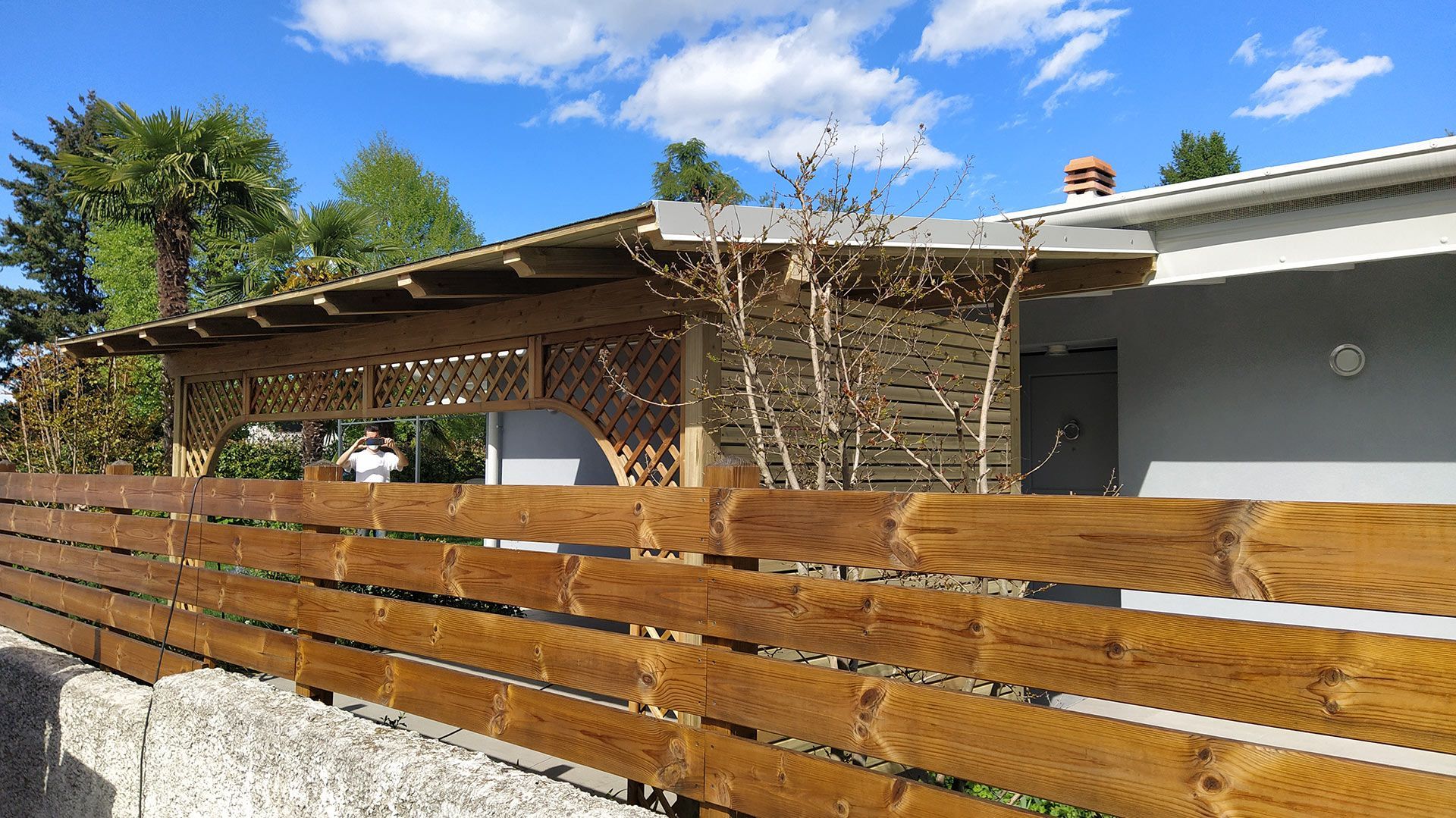 Recinzione a doghe di legno davanti a una casa con patio coperto e cielo azzurro.