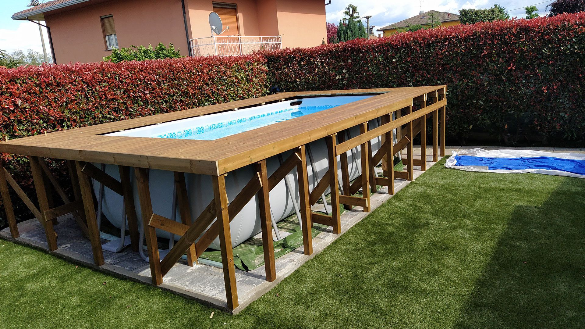 Piscina fuori terra circondata da una struttura in legno in un cortile con prato verde e siepi.