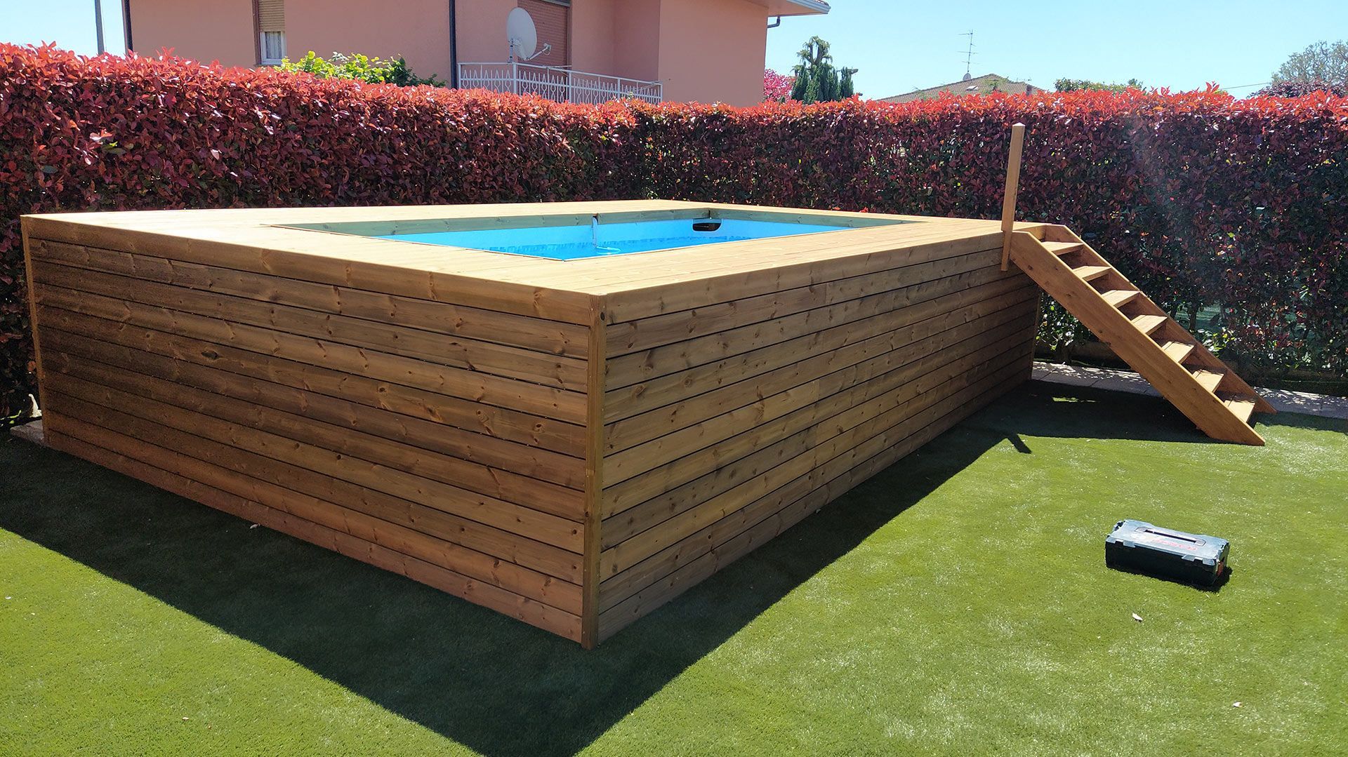 Piscina fuori terra in legno con acqua azzurra in un cortile, accanto a una piccola scala sulla destra.
