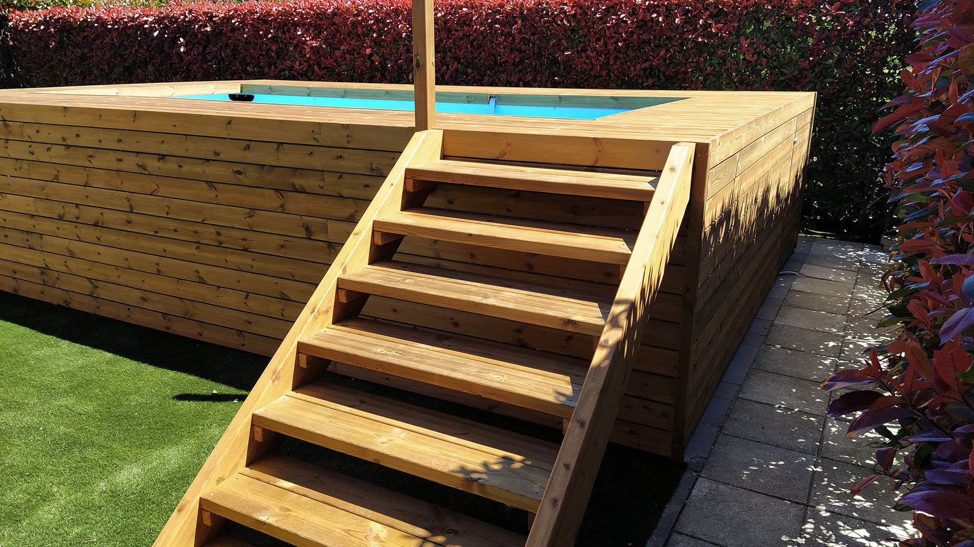 Terrazza in legno fuori terra con scalini, adiacente a un prato verde e a una siepe.