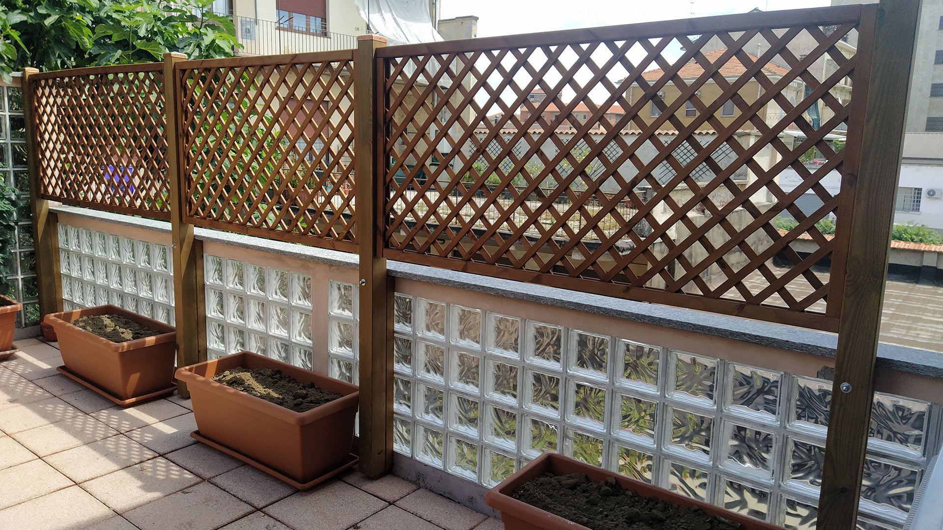 Recinzione a traliccio in legno con base in blocchi di vetro e fioriere lungo un patio piastrellato.