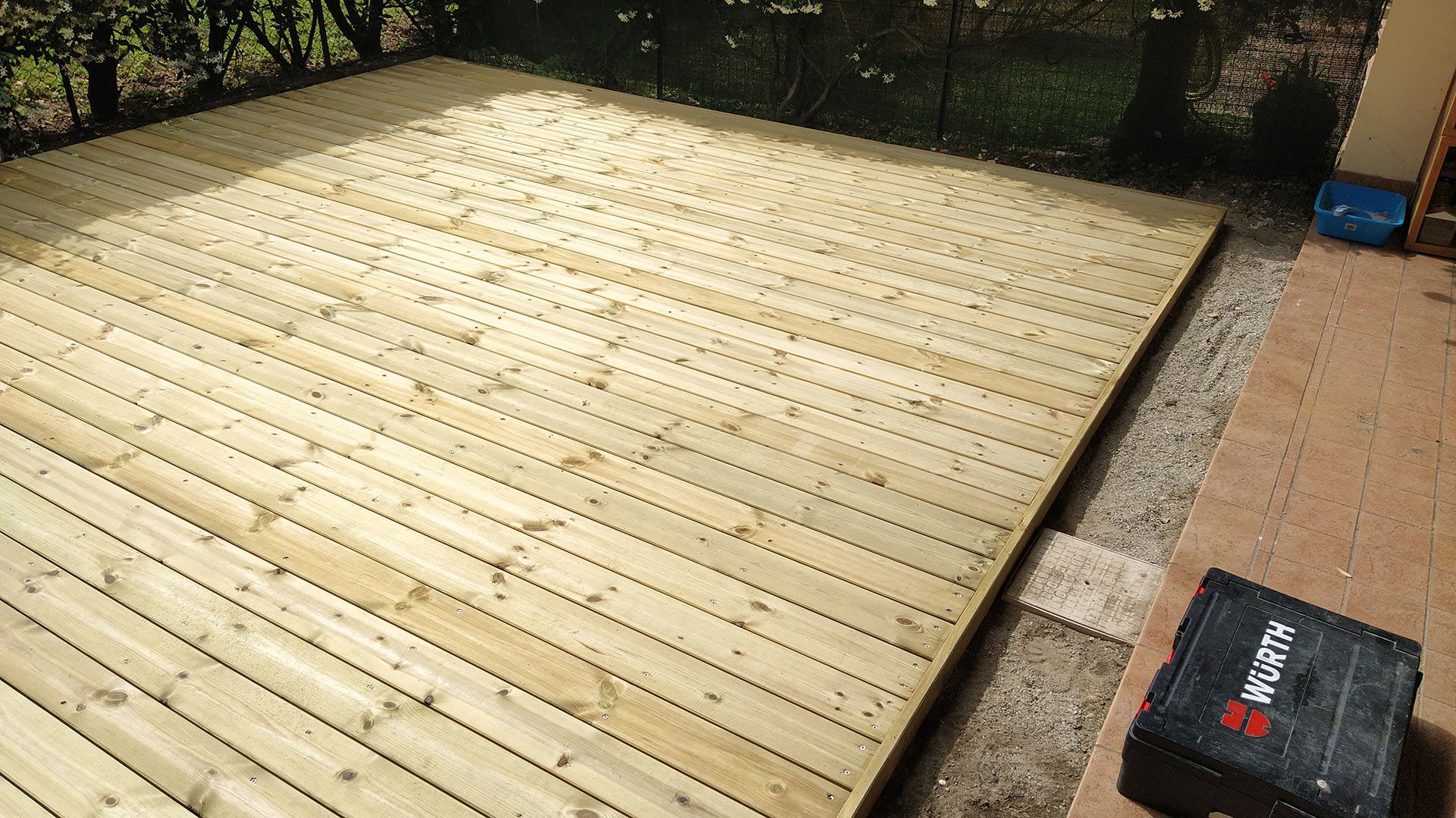 Terrazza in legno appena costruita in un cortile sul retro, adiacente a un terreno sterrato e lastricato con pietre da patio, circondata dal verde.