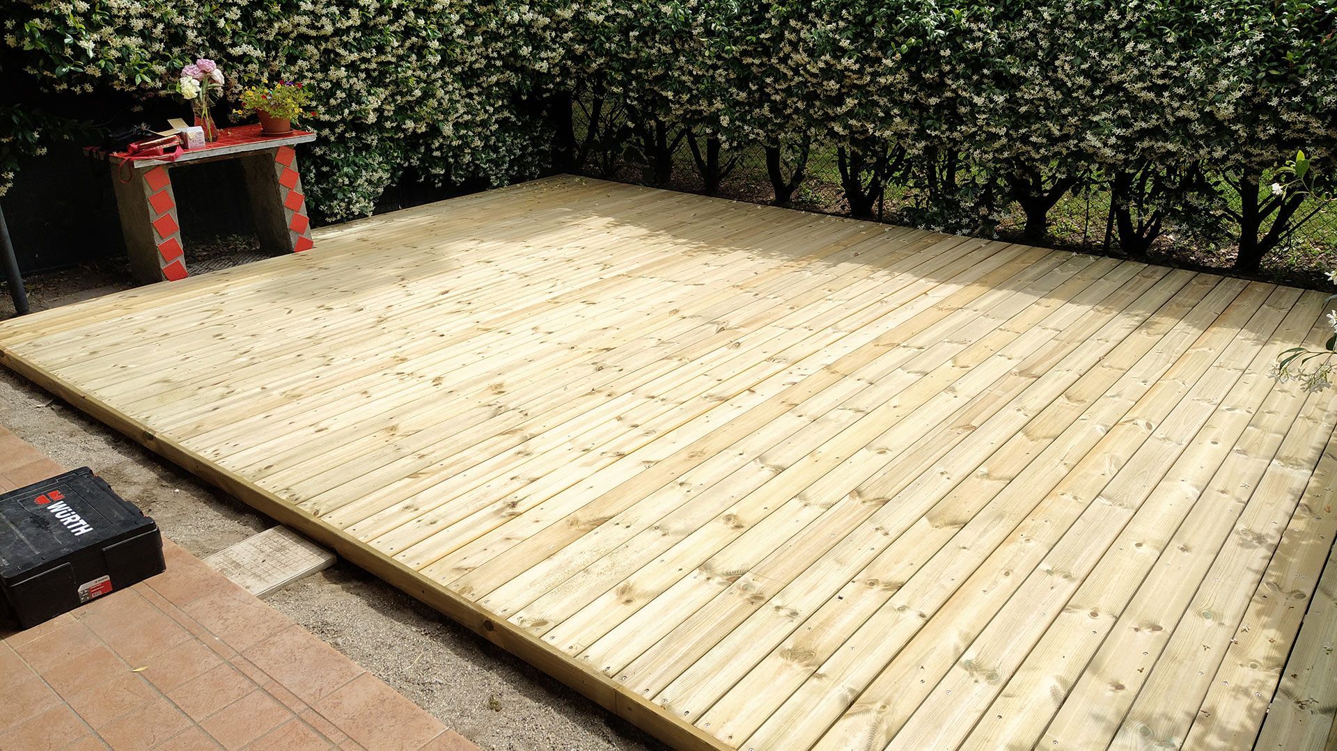 Terrazza in legno accanto a una siepe, con un tavolino rosso e una pianta in vaso in un angolo.