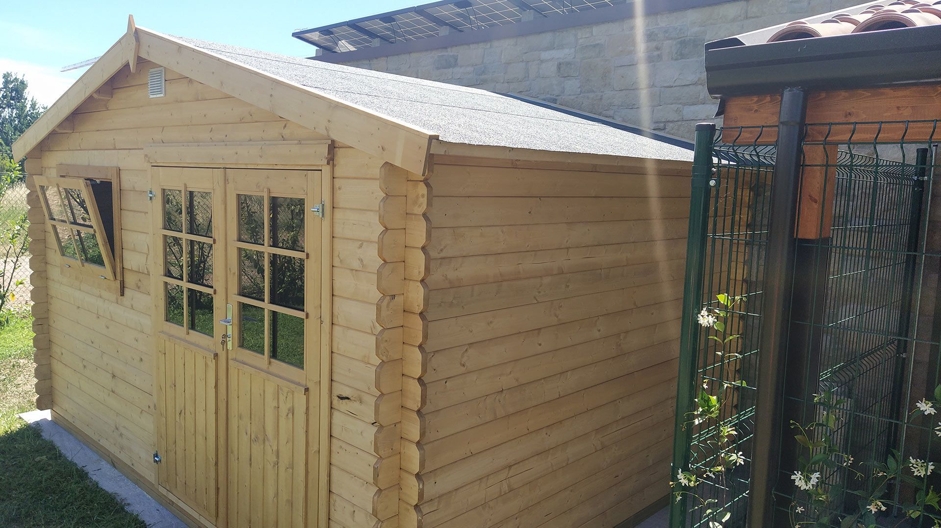 Casetta da giardino in legno con doppie porte accanto a una recinzione metallica nera in una giornata di sole.