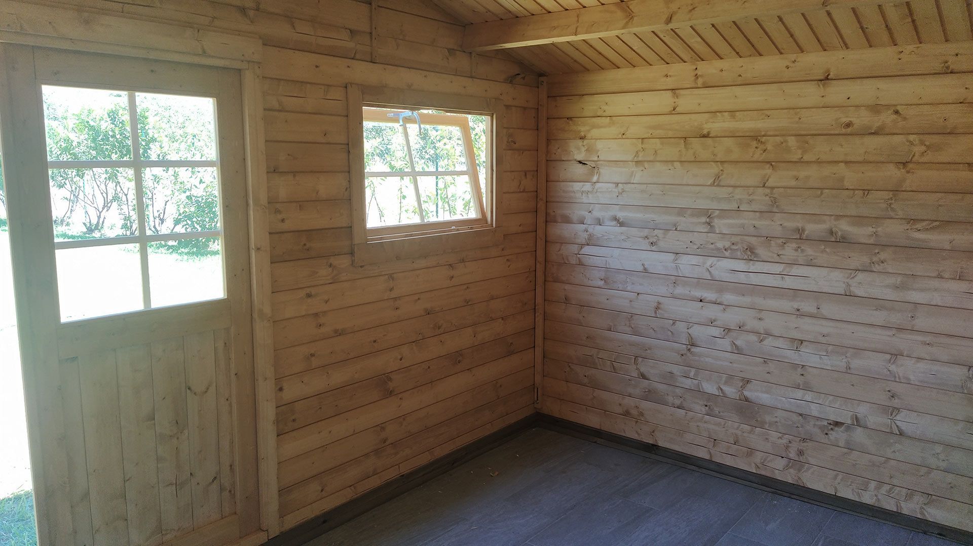 Stanza vuota rivestita in legno con porta finestrata e due piccole finestre che lasciano entrare la luce naturale.