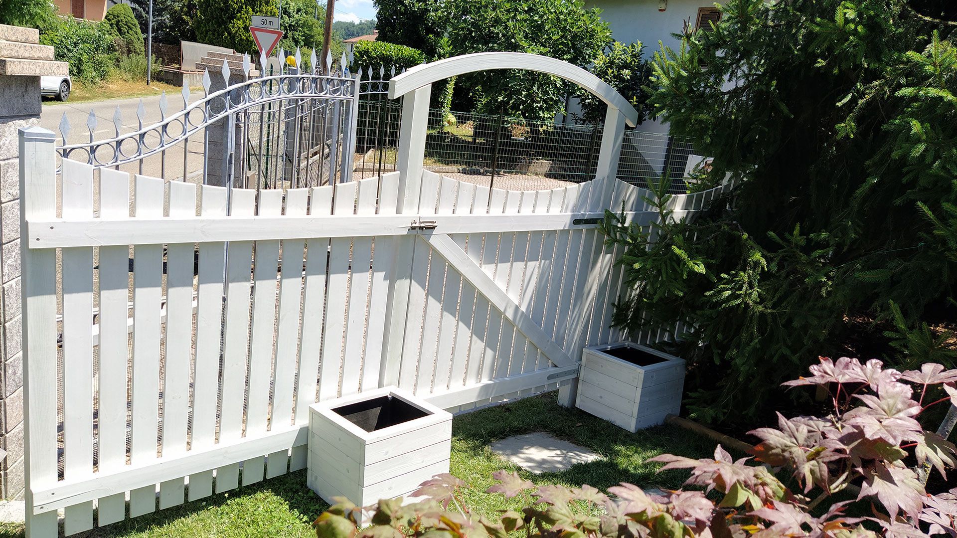 Recinzione bianca e cancello ad arco in un giardino soleggiato con piante in vaso e bordo in pietra.