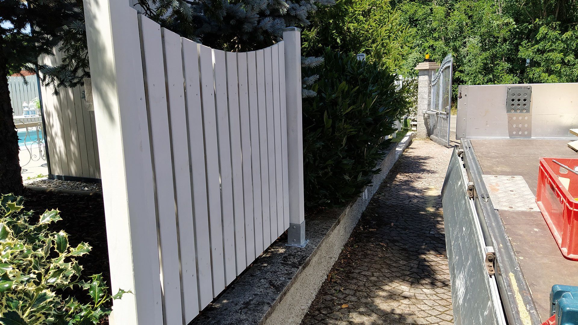 Stretto passaggio esterno accanto a una recinzione a doghe bianche, con alberi e una cassa rossa sulla destra.