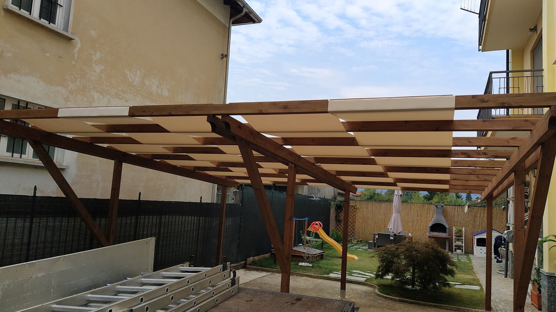 Pergola in legno in costruzione tra due case, con attrezzi e materiali nel cortile sul retro.