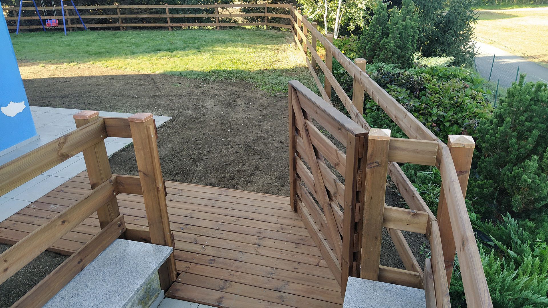 Rampa e scalini in legno che conducono a un cortile recintato con prato e alberi.
