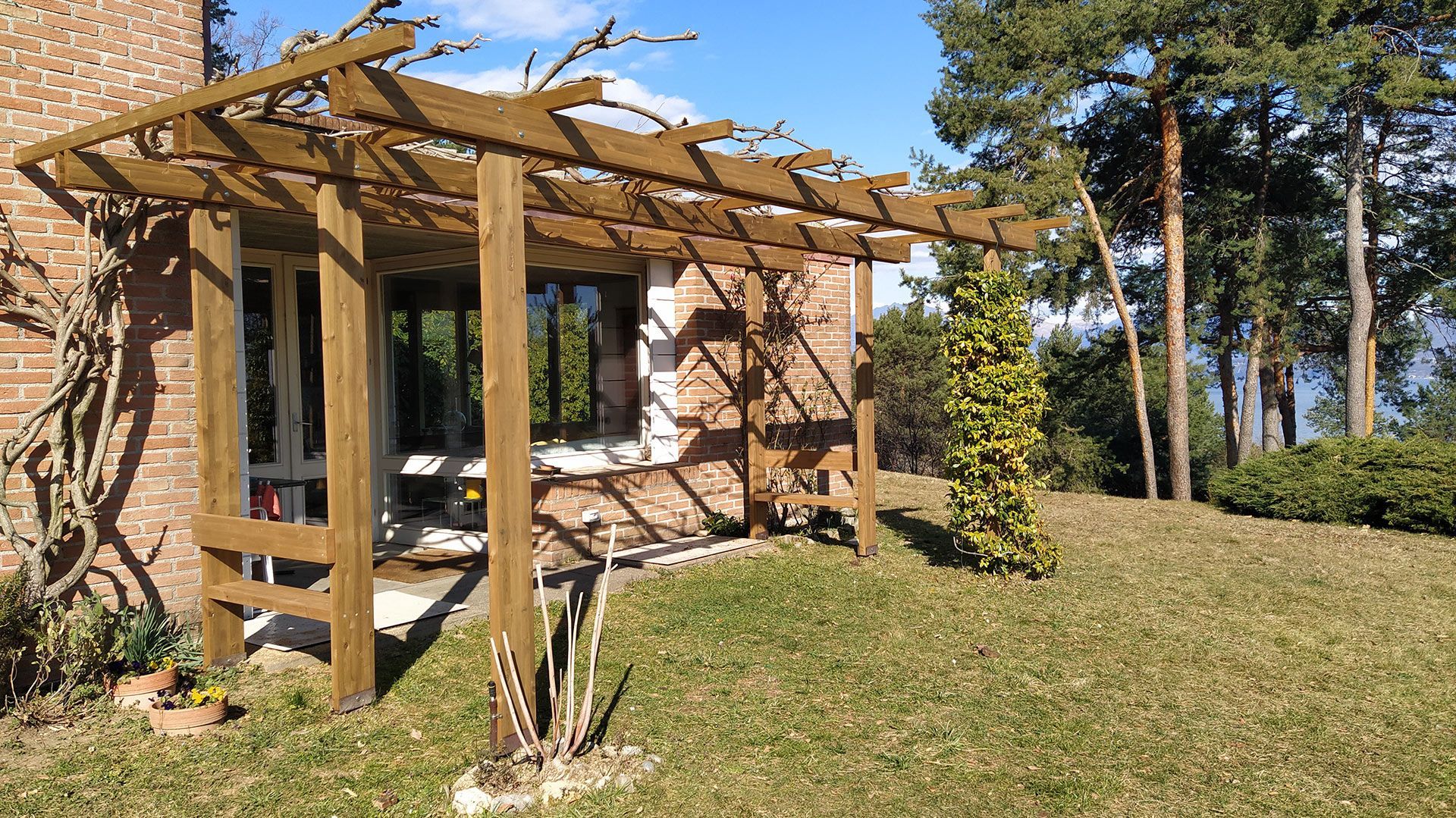 Pergola in legno accanto a una casa in mattoni in un soleggiato giardino erboso con alti pini.