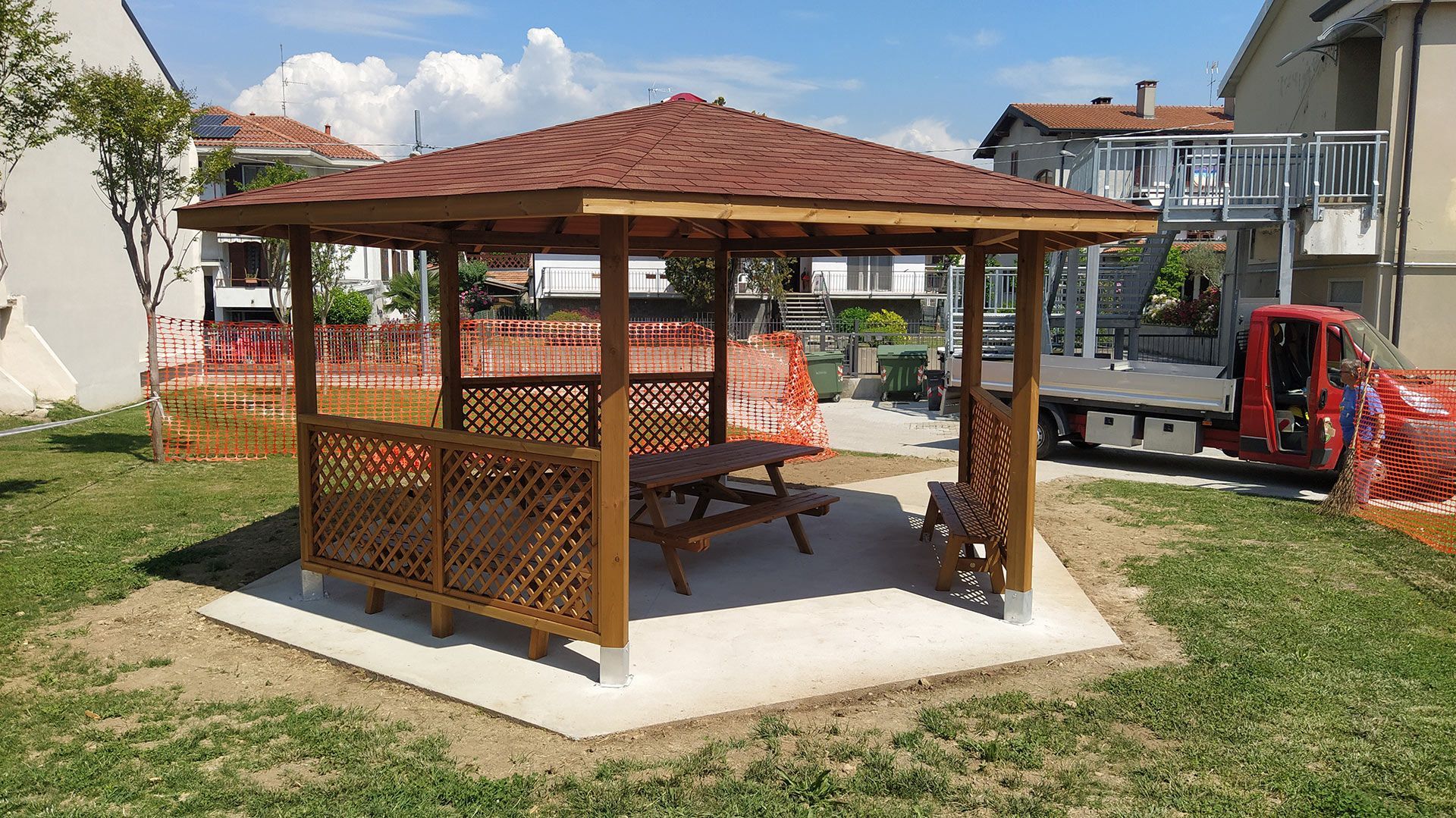 Gazebo in legno con panchine su una piattaforma di cemento in una zona esterna soleggiata, con edifici e attrezzature nelle vicinanze.