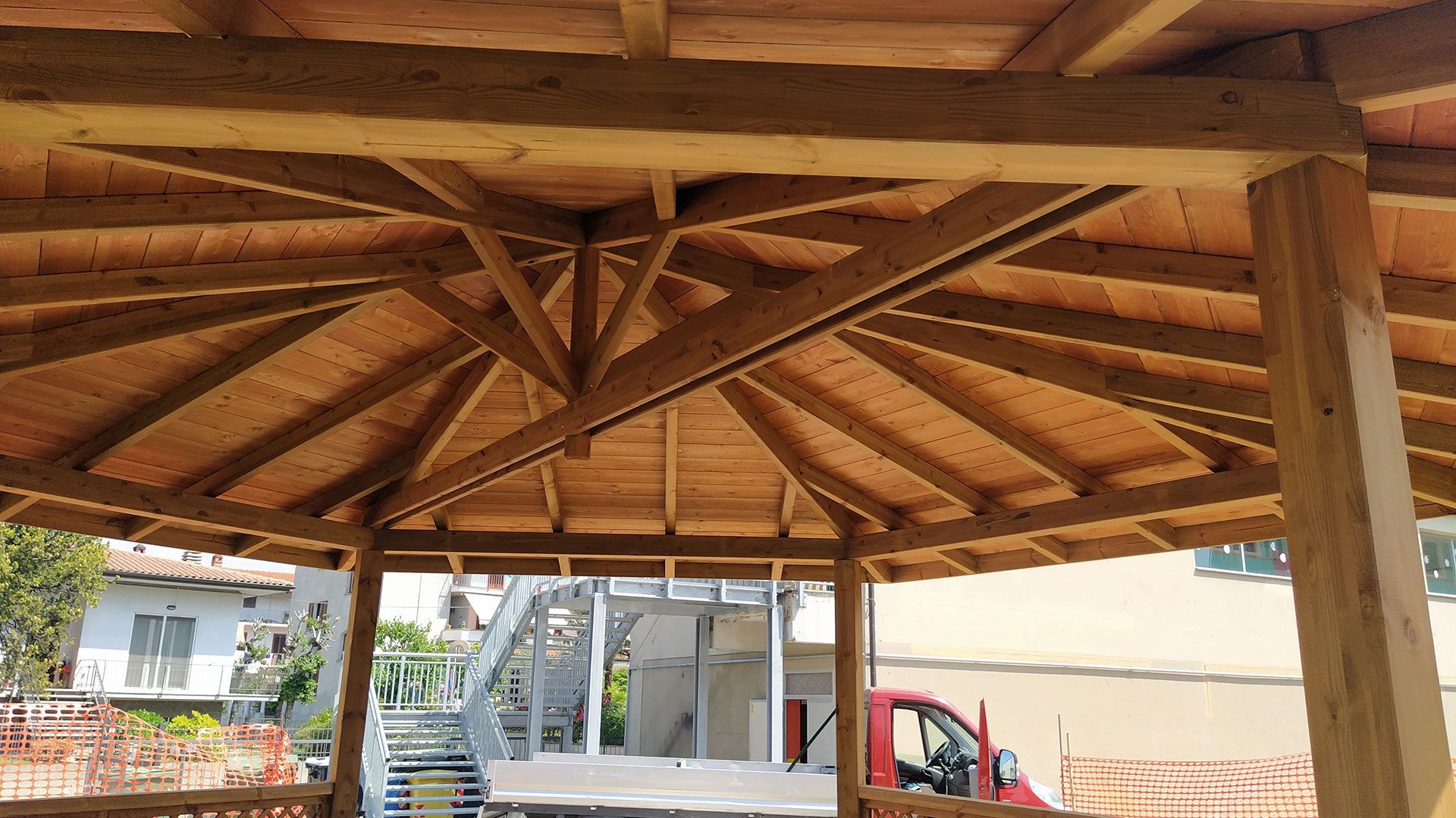 Struttura del tetto di un padiglione in legno con travi a vista sopra un vialetto, con un camion bianco e un'auto rossa sottostanti.
