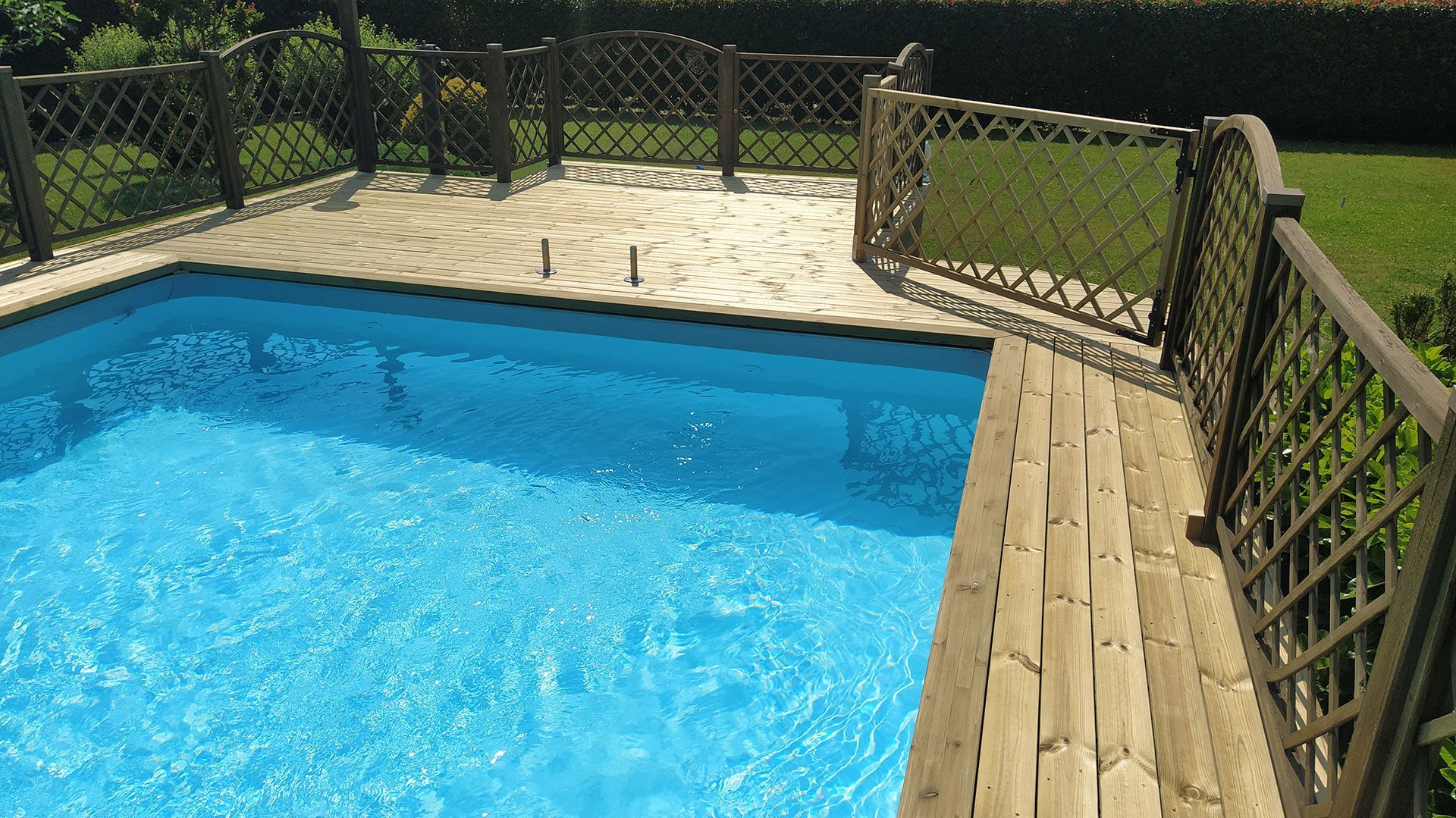 Piscina in giardino con acqua azzurra, pavimentazione in pietra e recinzione in legno in una giornata di sole.