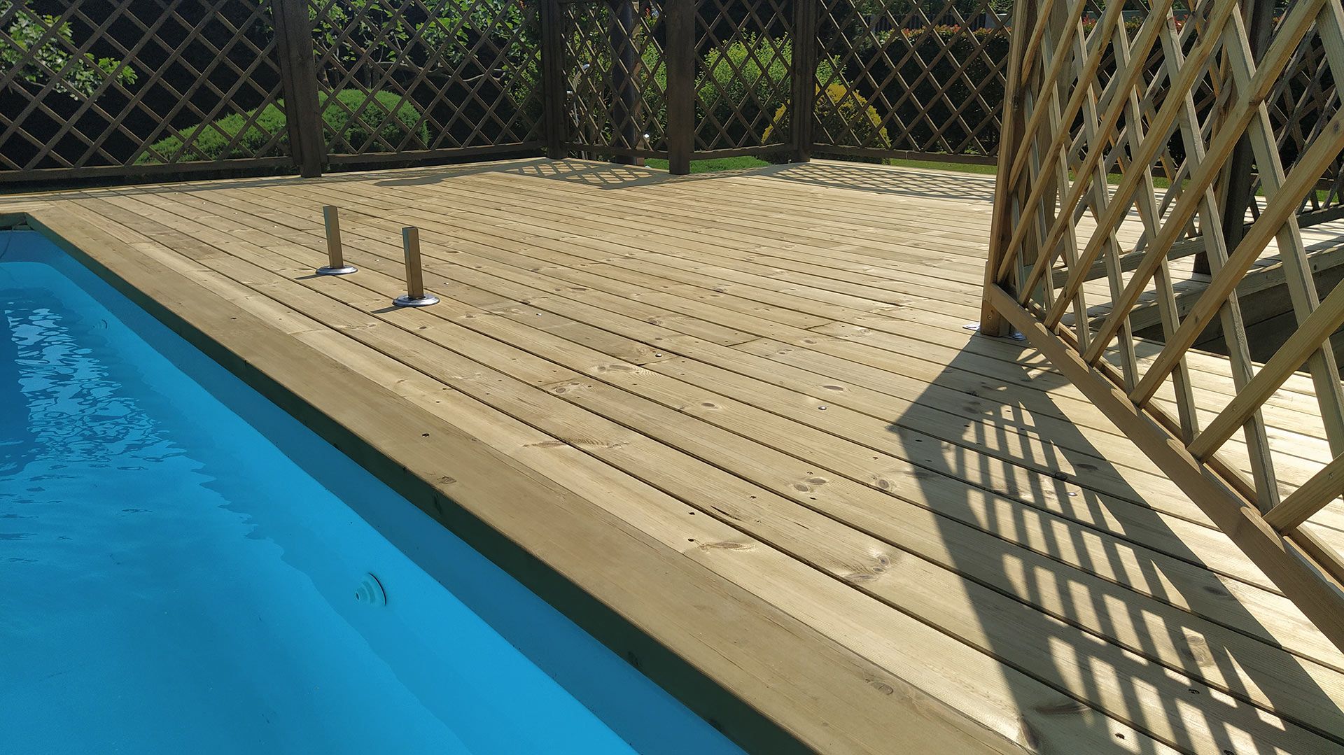 Terrazza in legno a bordo piscina, accanto a una piscina blu, con recinzione a traliccio e recinzione a rete metallica, illuminata dal sole.
