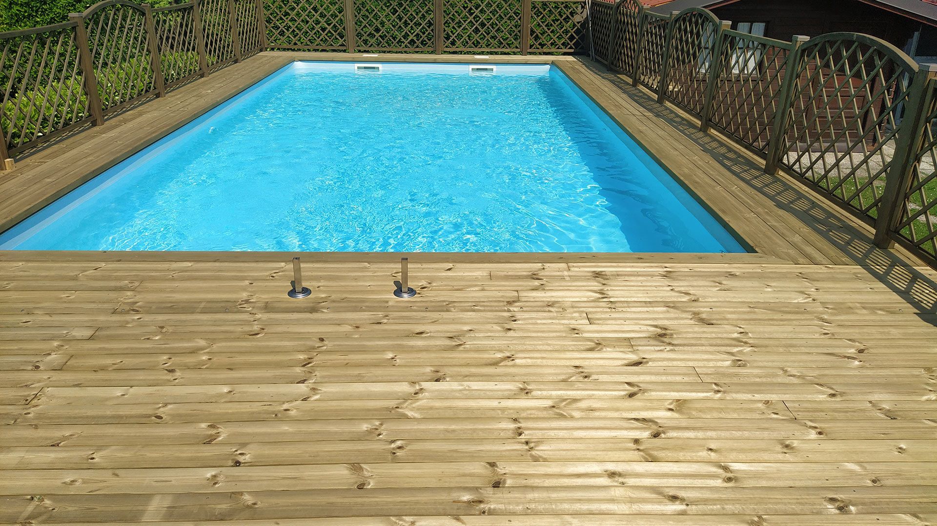 Piscina in giardino con terrazza e recinzione in legno, esposta alla luce del sole.