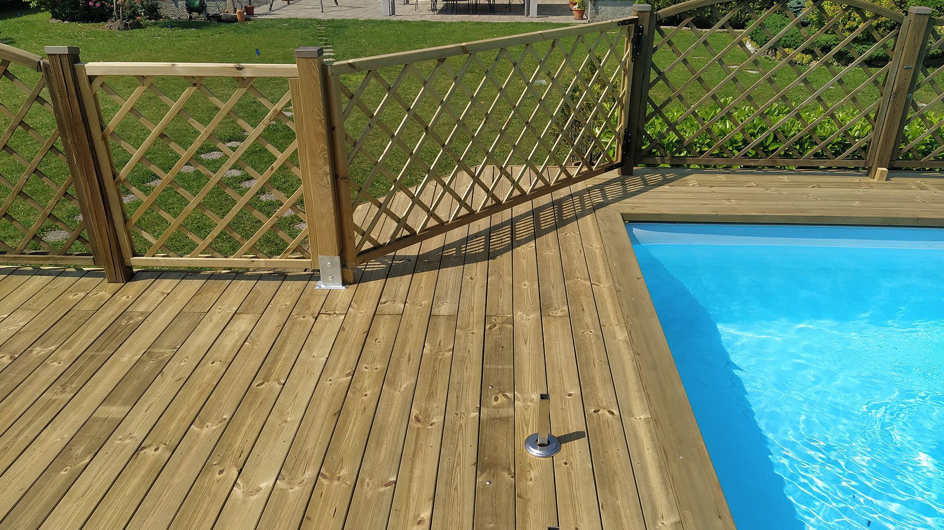 Terrazza in legno accanto a una piscina di un blu brillante, con una recinzione a traliccio e un prato alle spalle.