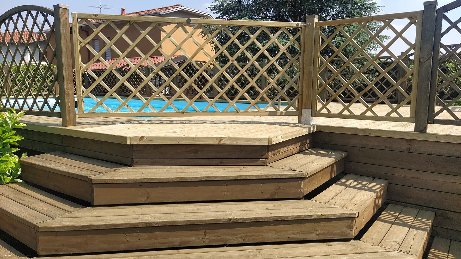Terrazza in legno sul retro della casa con scalini e ringhiera a traliccio, affacciata su una piscina blu in una giornata di sole.
