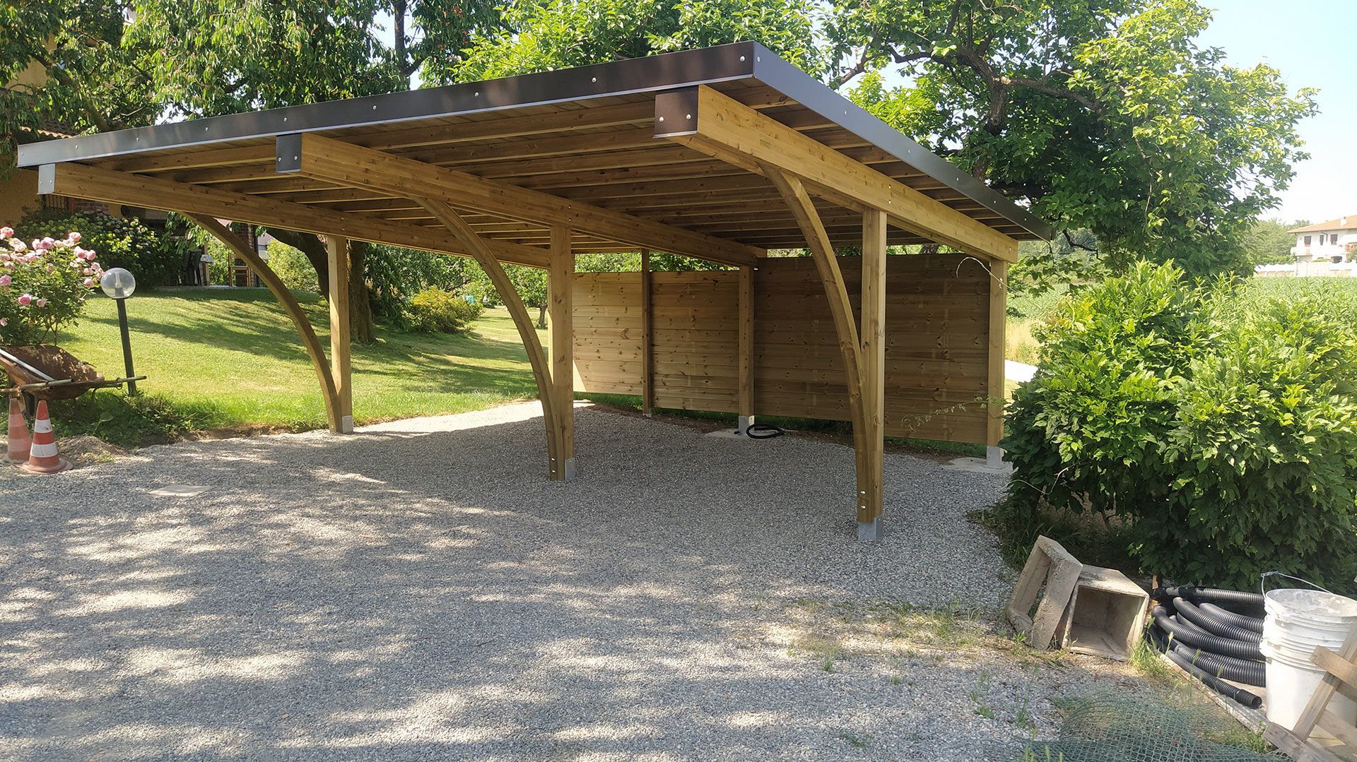 Posto auto coperto in legno con tetto spiovente su un vialetto di ghiaia, circondato da alberi e vegetazione.