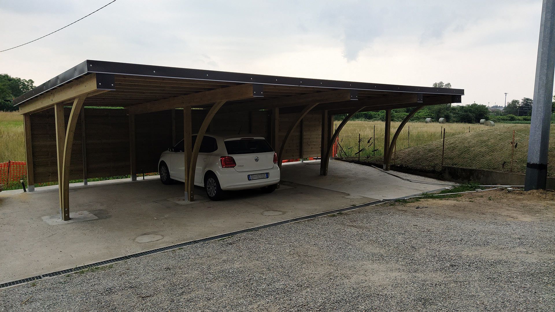 Auto bianca parcheggiata sotto una tettoia di legno a lato di una strada sterrata in una zona rurale.