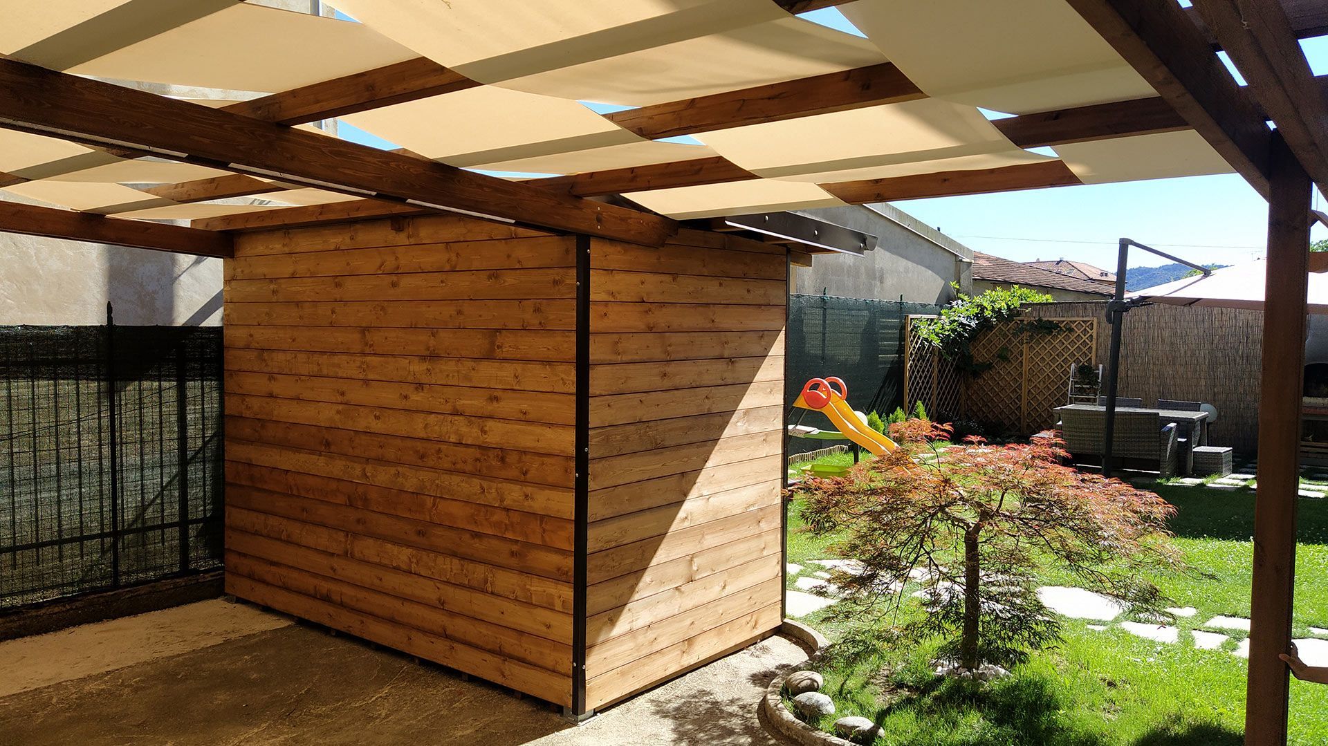 Casetta in legno sotto un pergolato a doghe in un soleggiato giardino sul retro, con prato e fiori.
