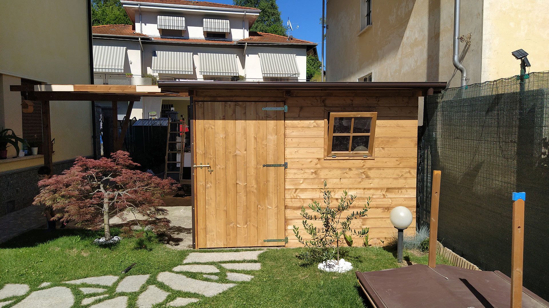 Casetta da giardino in legno con doppie porte e finestra, situata in un piccolo cortile sul retro, con vialetto in pietra e prato.