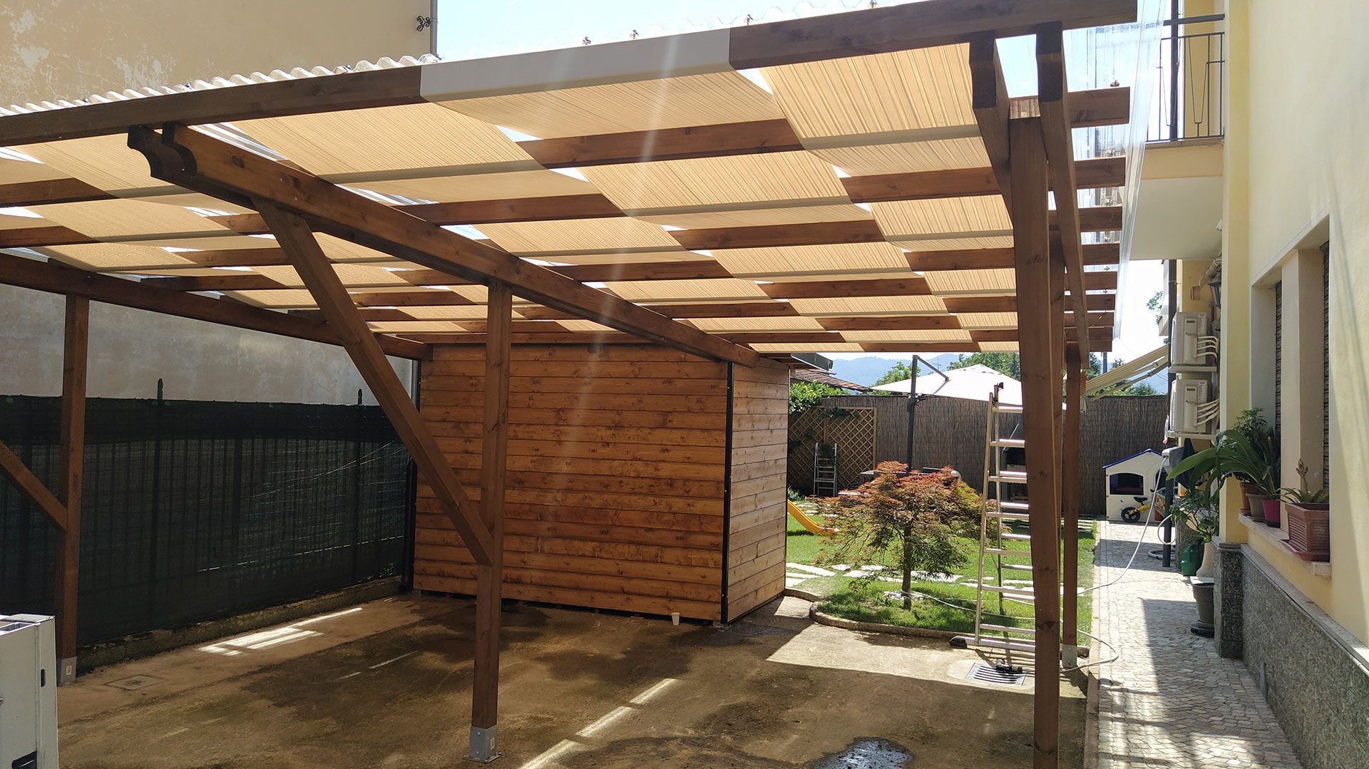 Pergola in legno sopra un patio pavimentato accanto a una casa, con un piccolo giardino e una recinzione sullo sfondo.