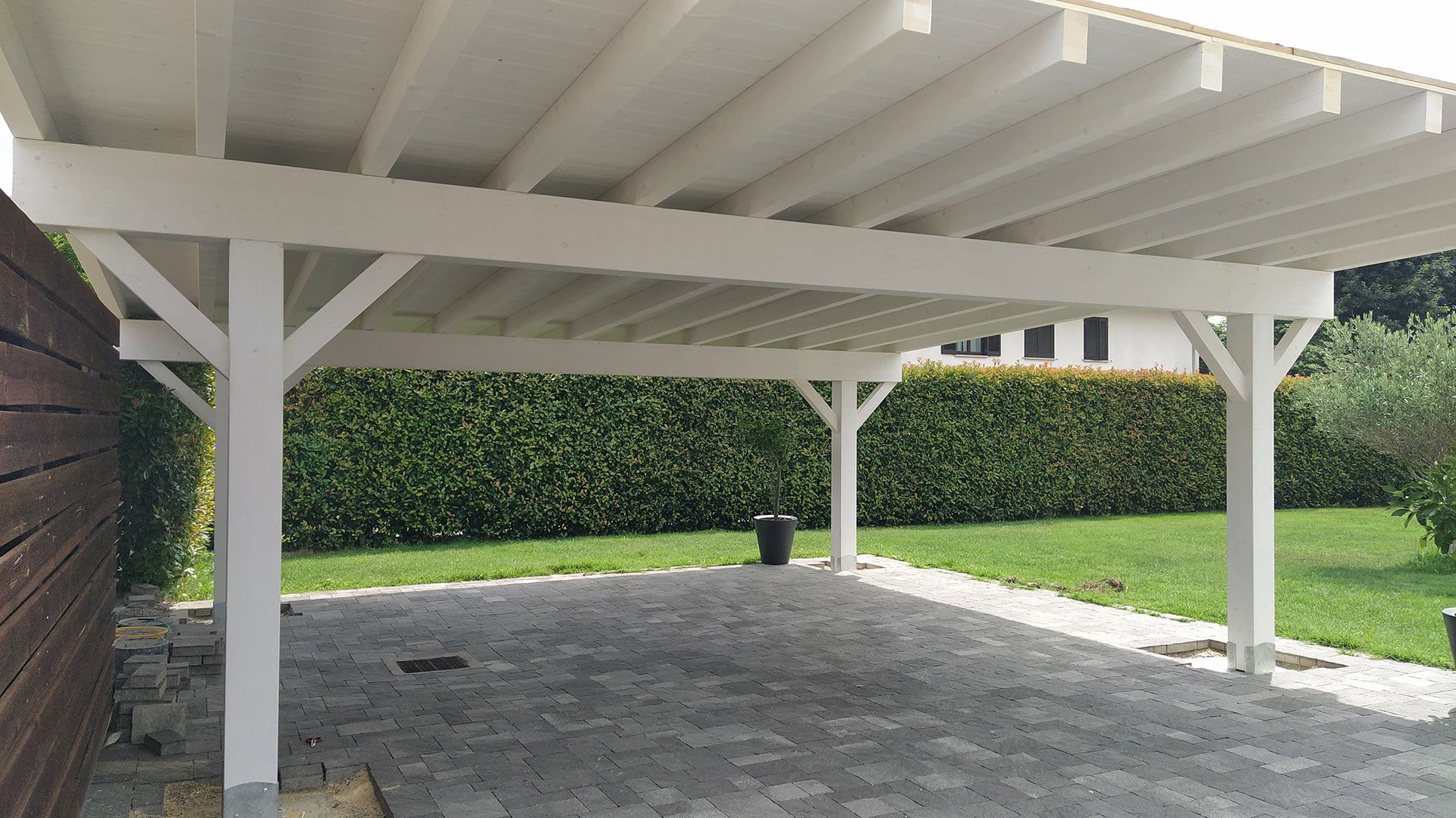 Area di parcheggio esterna coperta con pali bianchi, pavimentazione e siepi sullo sfondo.