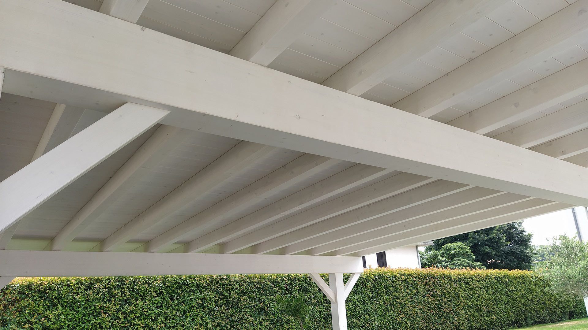 Tettoia bianca a pergola con travi inclinate sopra una siepe verde in un ambiente esterno