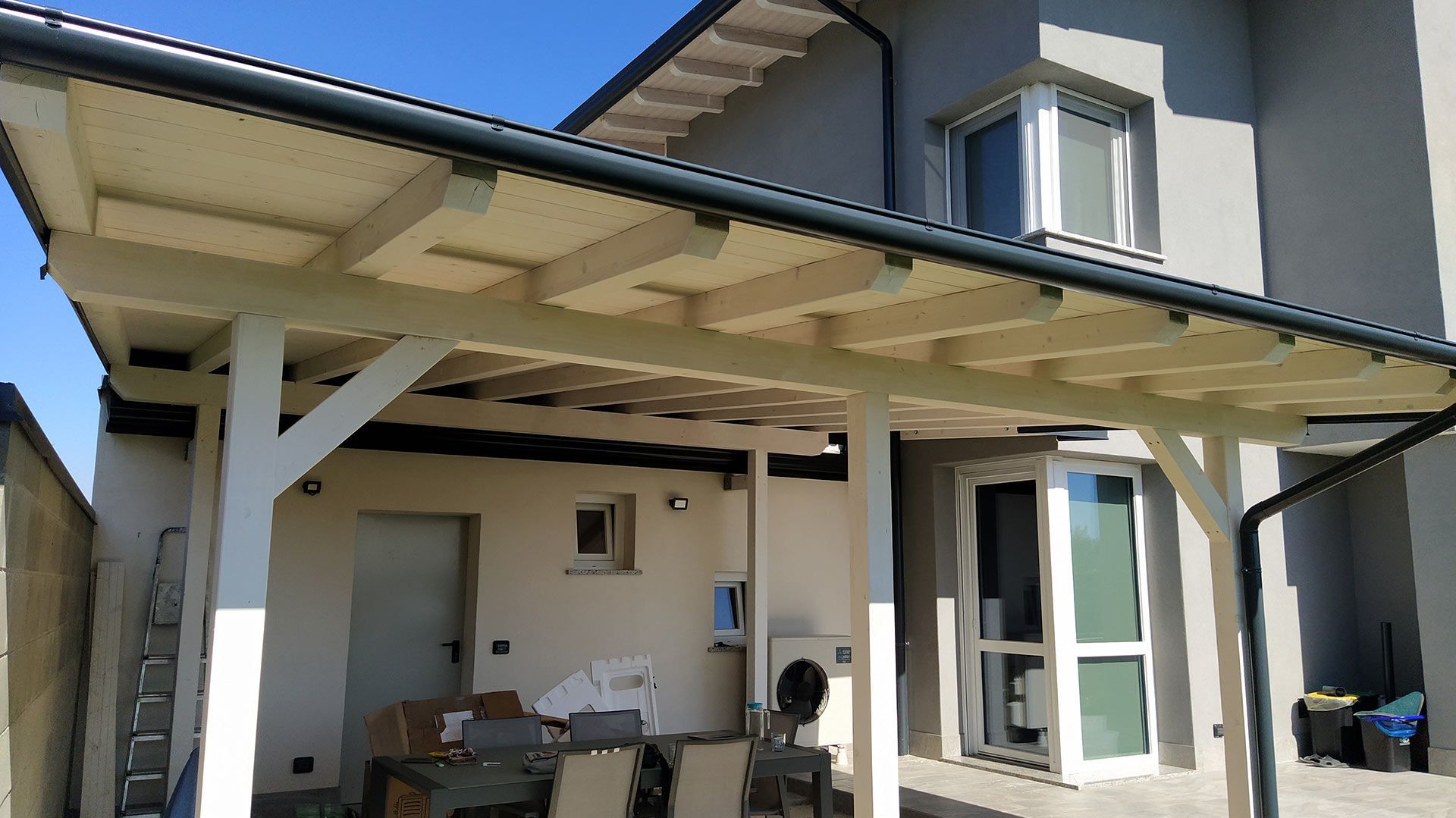 Patio coperto annesso a una casa, con pali di sostegno bianchi, rivestimento esterno color beige e mobili da esterno.