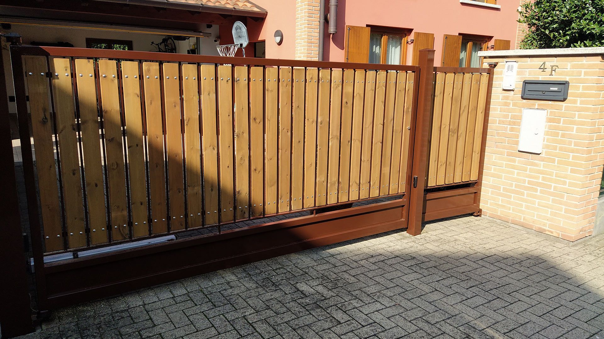 Cancello carrabile in legno con pali in metallo nero accanto a un muro di mattoni e a un cortile pavimentato, di fronte a una casa