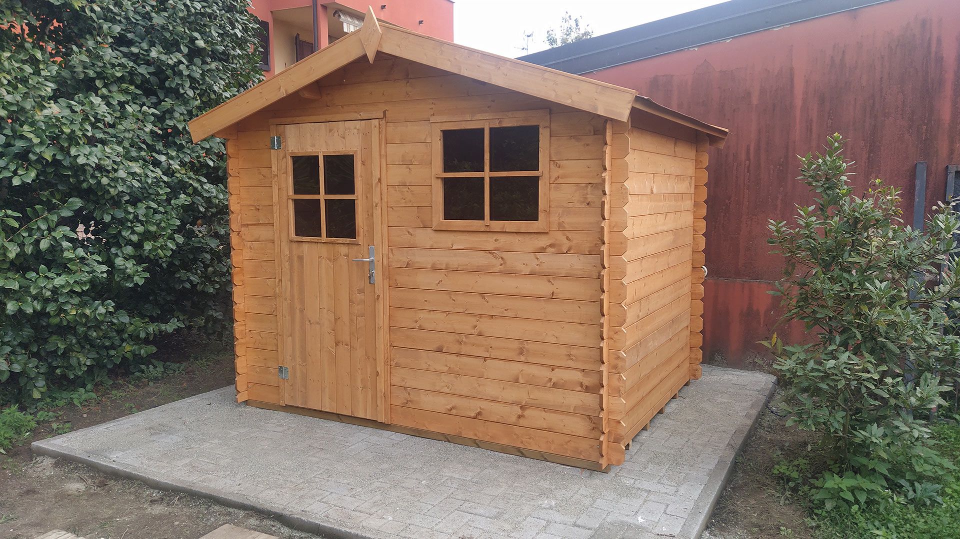 Piccola casetta da giardino in legno con due finestre e una porta, su una base di cemento, accanto a una siepe e a una recinzione.