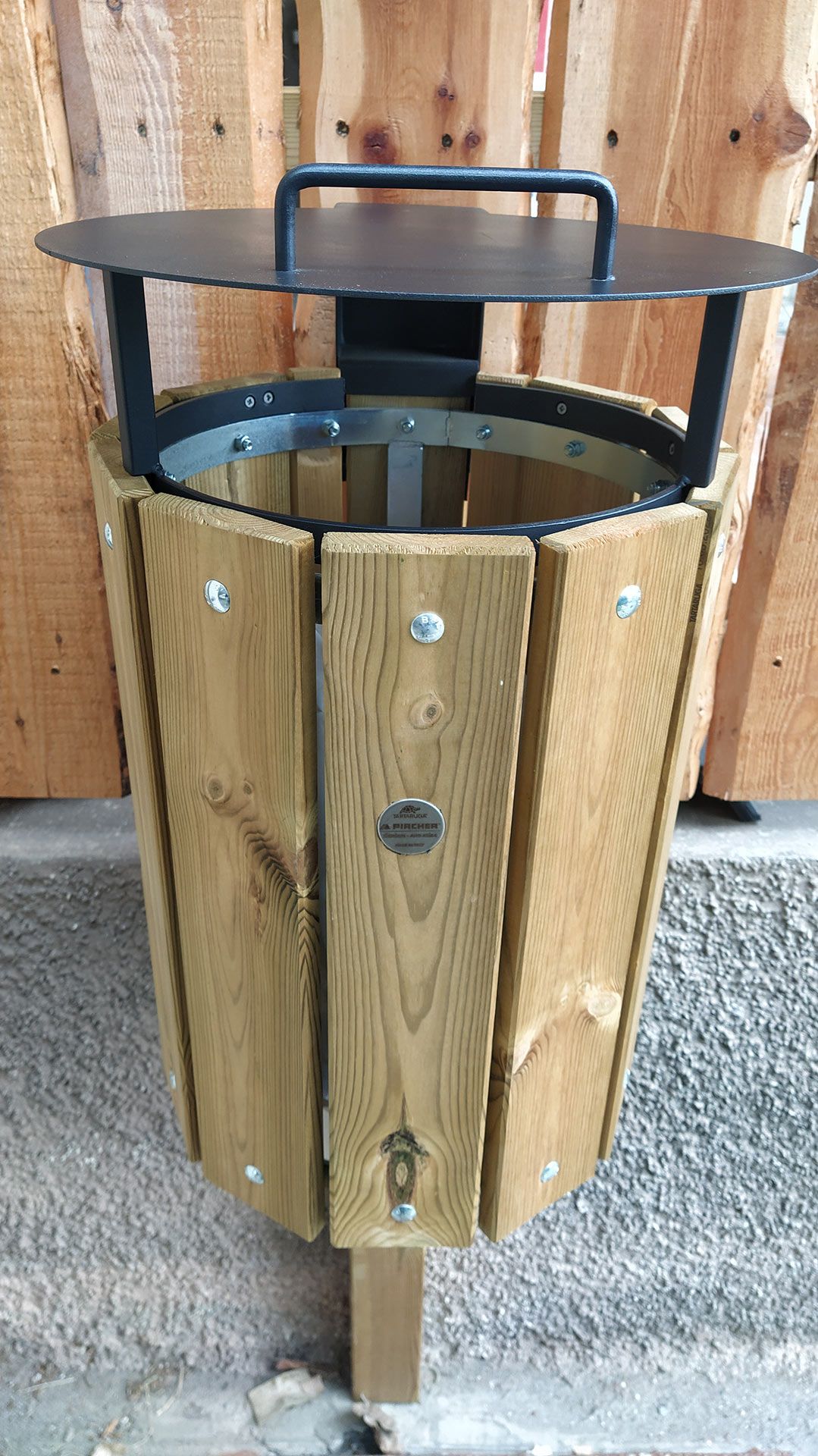 Pattumiera da esterno in legno con coperchio e maniglia superiore in metallo nero, fissata a una recinzione in legno.