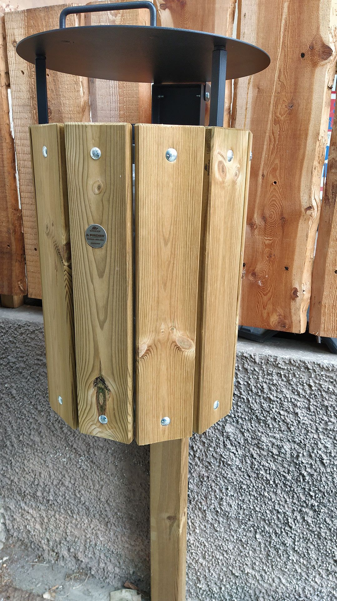 Cestino per rifiuti da esterno in legno, addossato a un muro di legno invecchiato e a una sporgenza in pietra.