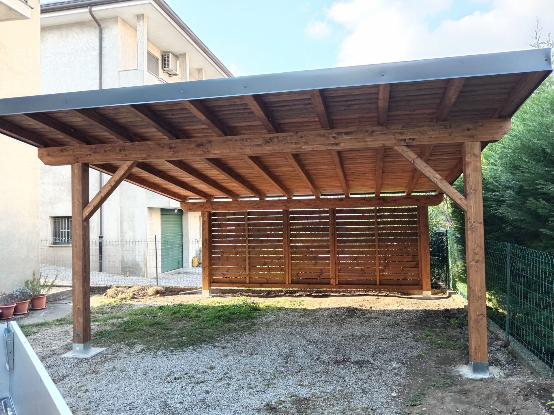 Posto auto coperto in legno con tetto spiovente, adiacente a una casa e situato sopra un vialetto di ghiaia.