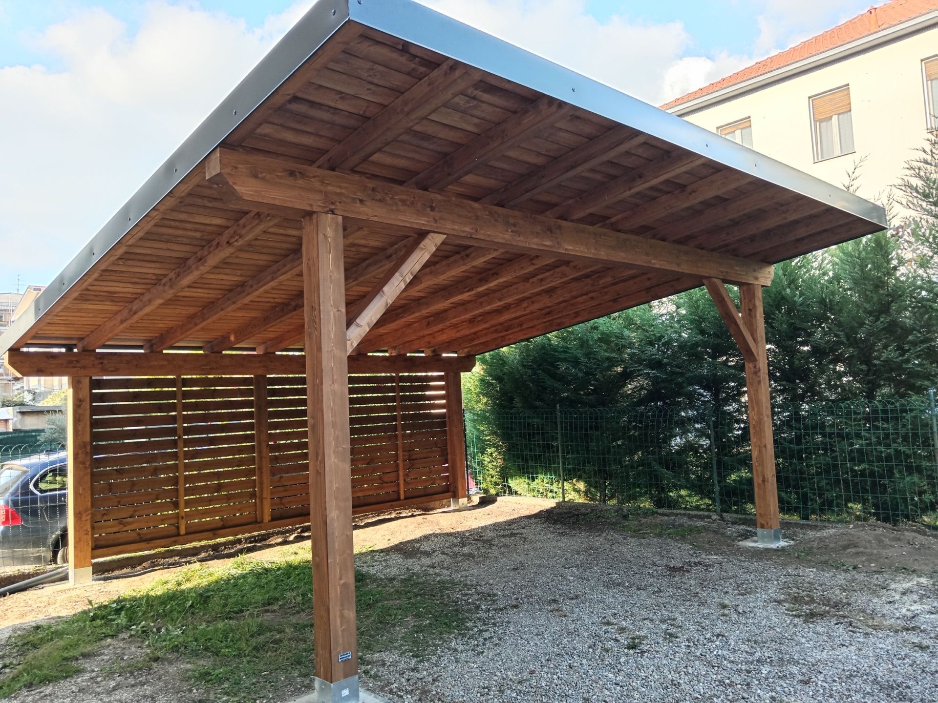 Posto auto coperto in legno con tetto spiovente su ghiaia accanto a una casa e a una recinzione