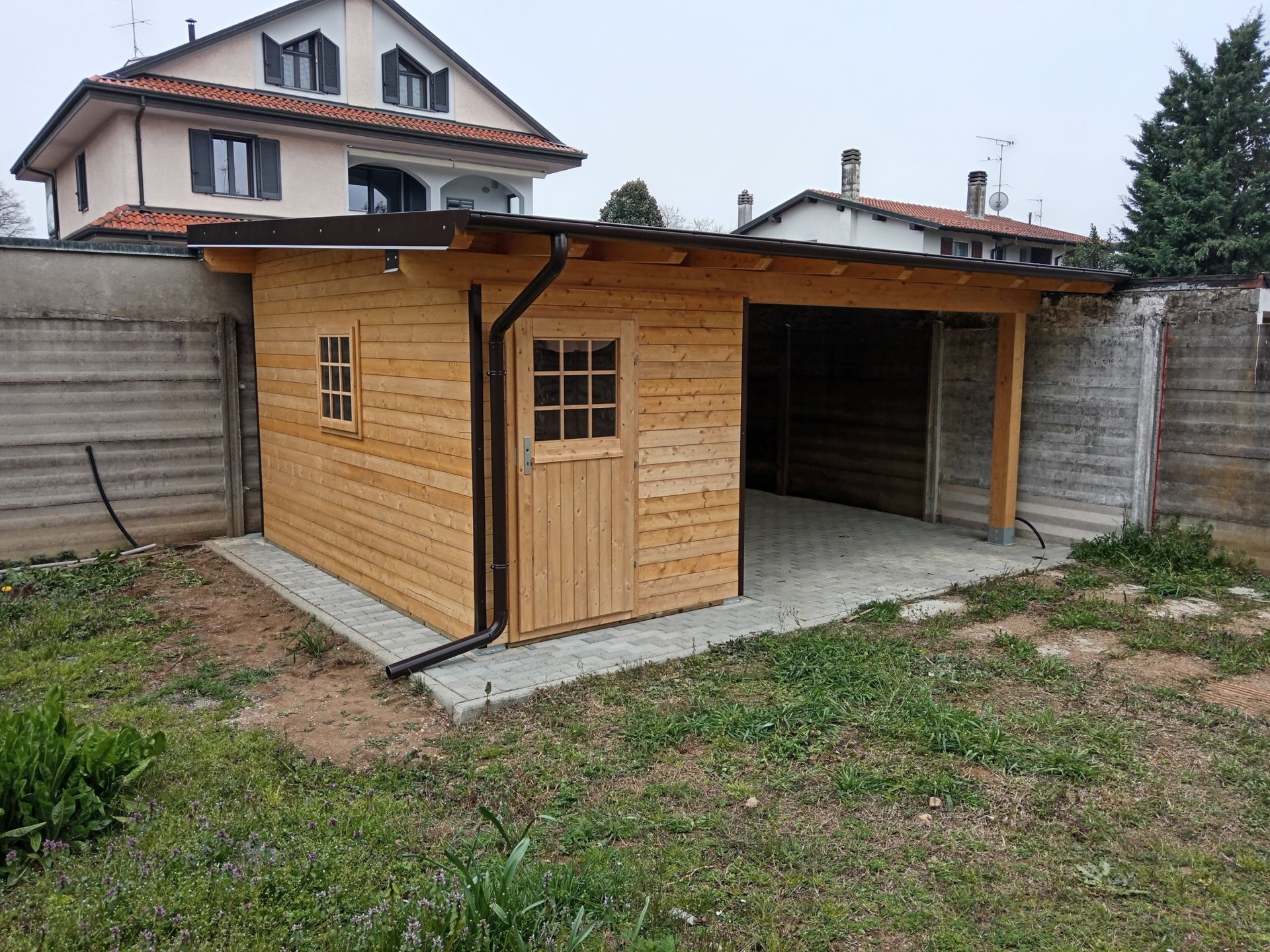 Garage e capanno in legno nel cortile sul retro, con posto auto coperto aperto, situati accanto a un muro di cemento e a un prato.