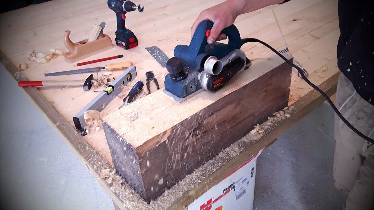 In un'officina, una persona utilizza una pialla elettrica portatile su una trave di legno.
