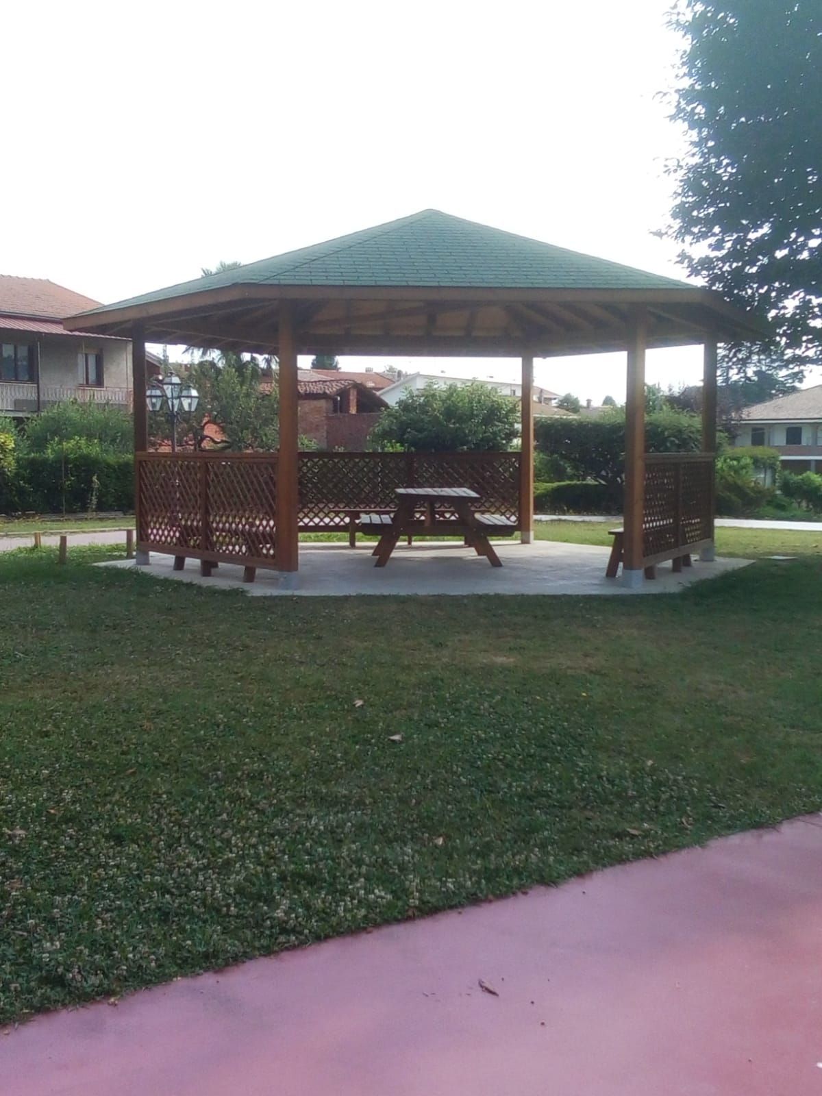 Gazebo aperto con tetto verde in un parco erboso, con panchine e un sentiero lastricato nelle vicinanze.