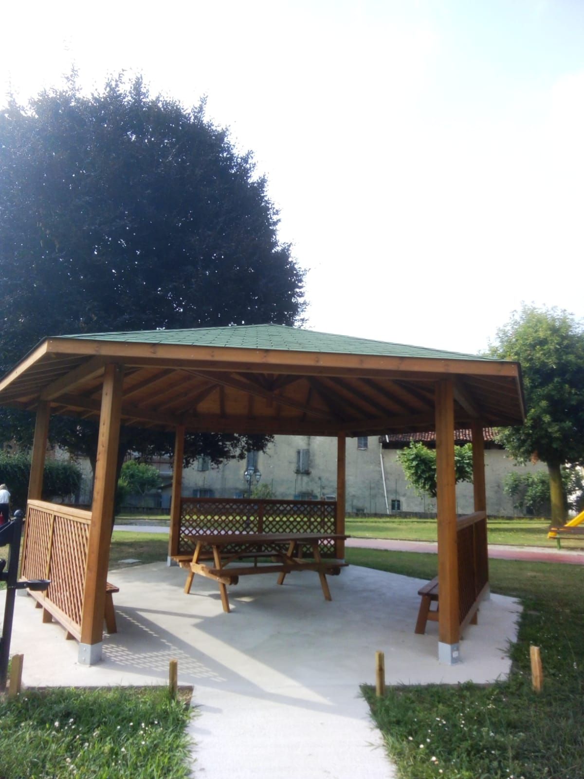 Gazebo in legno con tetto verde e panchina in un parco in una giornata di sole.