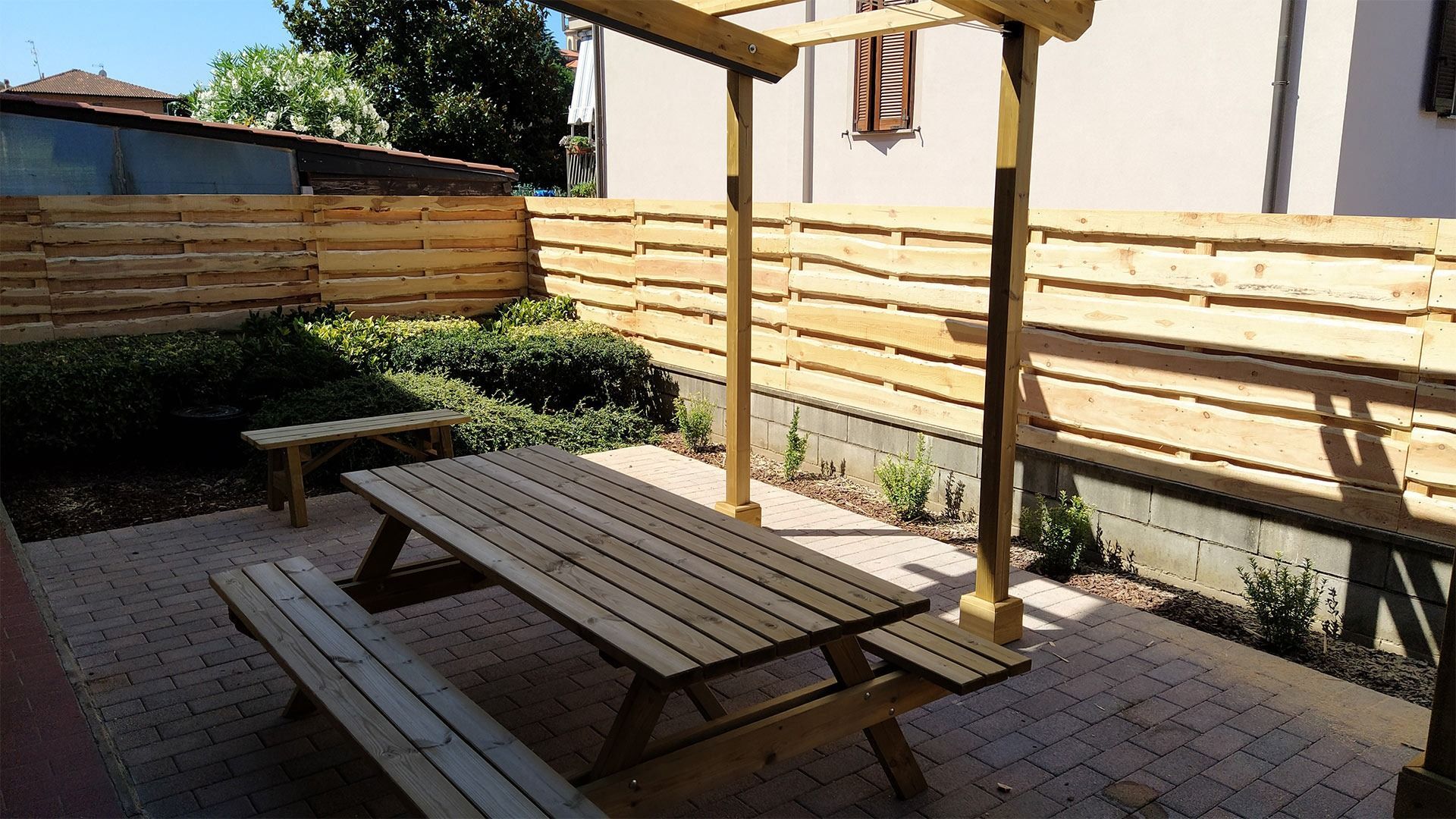 Patio esterno con tavolo da picnic in legno, panchine e pergolato accanto a un cortile recintato e soleggiato.