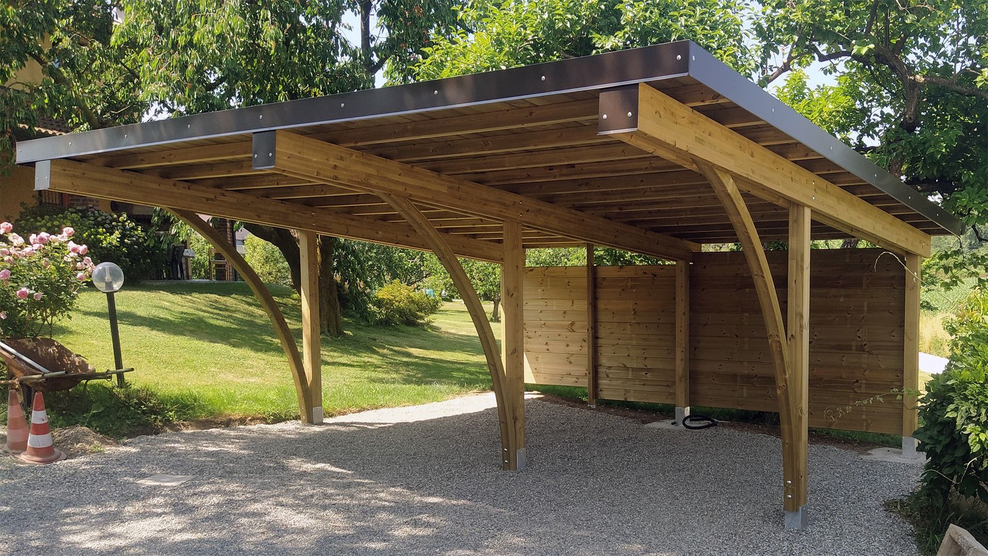 Posto auto coperto in legno aperto su un lato, con tetto spiovente, su un vialetto di ghiaia, circondato da alberi ed erba.