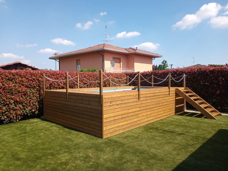 Terrazza rialzata in legno con ringhiera e scalini in corda, immersa nel verde di un cortile, con una casa e siepi sullo sfondo.