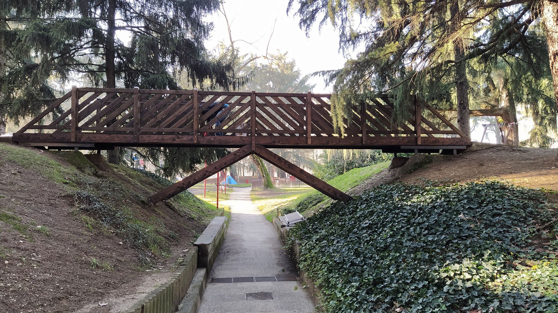 Una passerella di legno attraversa uno stretto sentiero di cemento all'interno di un parco paesaggistico, con argini ricoperti di edera e alberi.