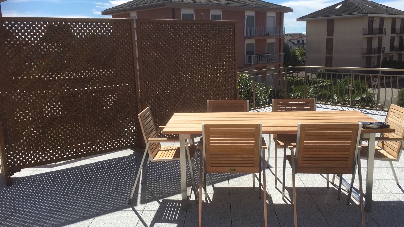Patio esterno con tavolo e sedie in legno accanto a una schermatura a traliccio, in pieno sole.