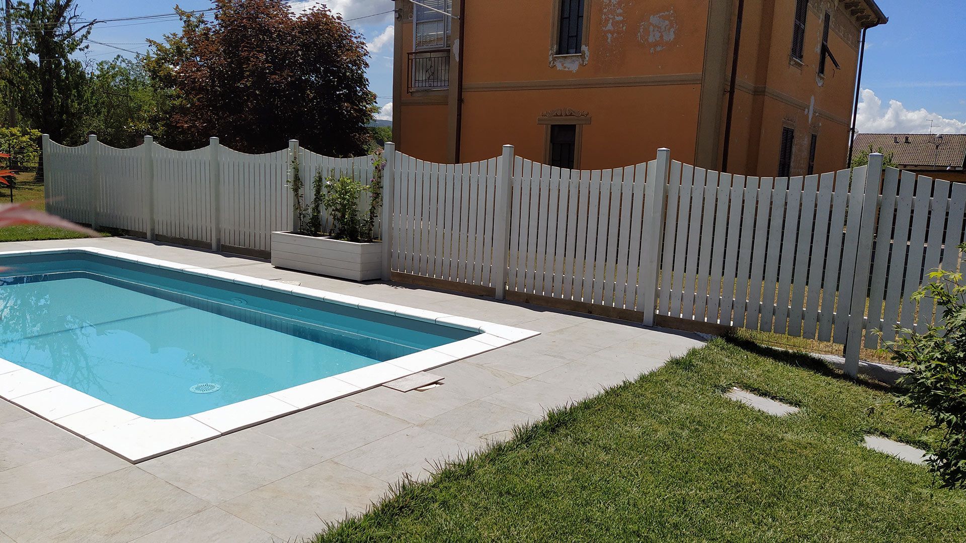 Piscina nel cortile sul retro, accanto a una staccionata bianca, con prato e casa arancione sullo sfondo.