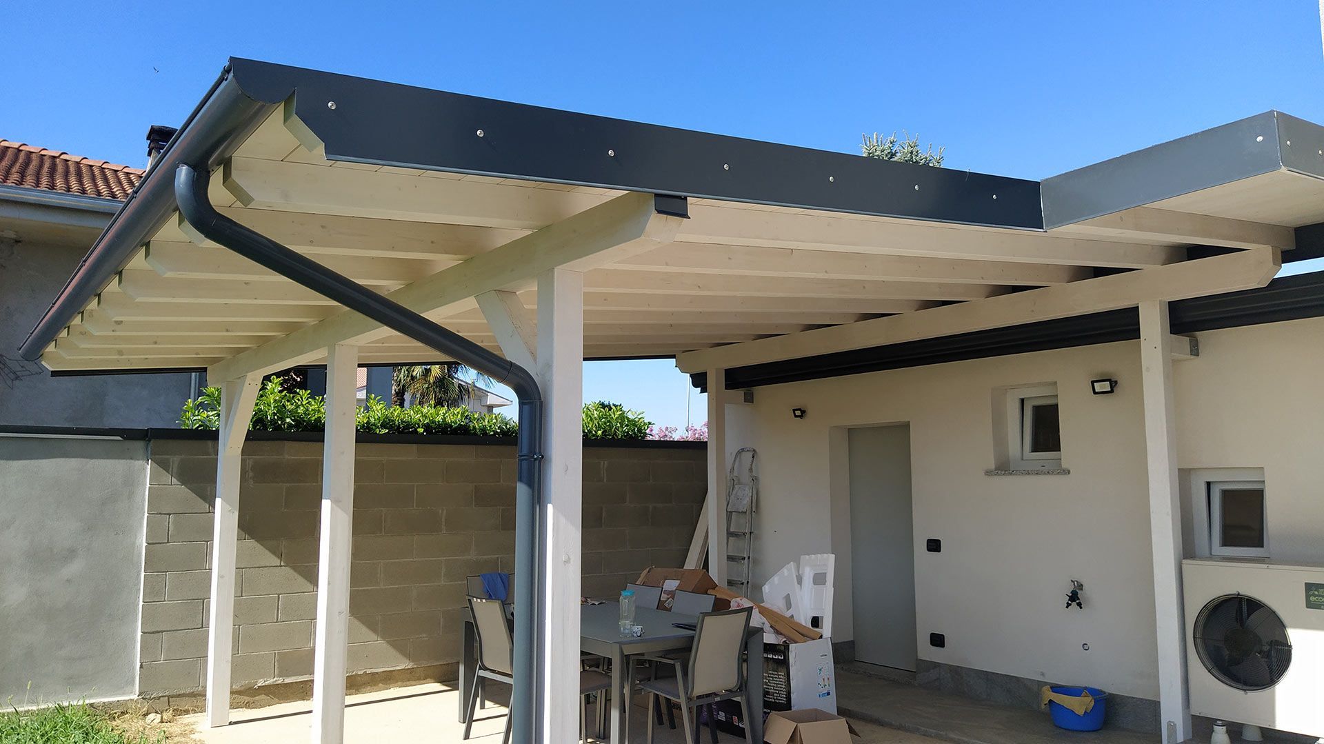 Patio coperto accanto a una casa bianca con tavolo, sedie e attrezzature da esterno sotto un cielo azzurro.