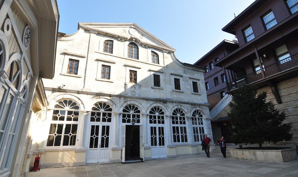 A large white building with arched windows is surrounded by smaller buildings