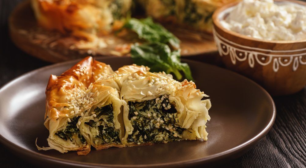 A slice of spinach pie is on a plate next to a bowl of sour cream.